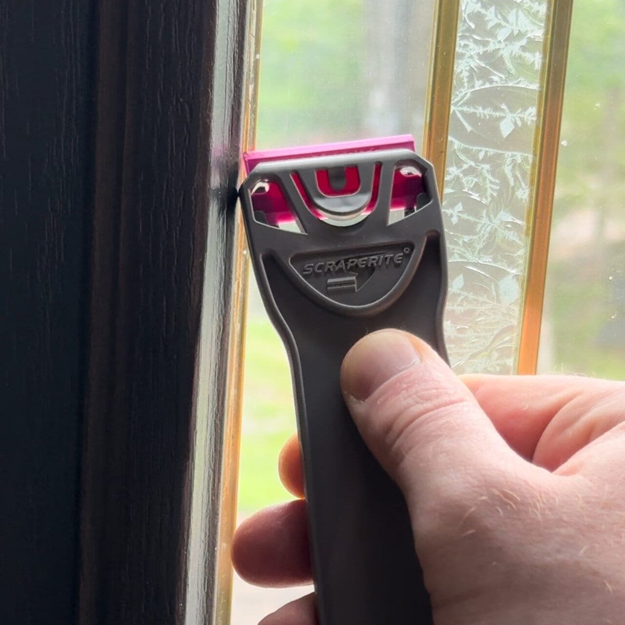 Hand holding Scraperite Pro Magenta Blade scraper against a window frame.