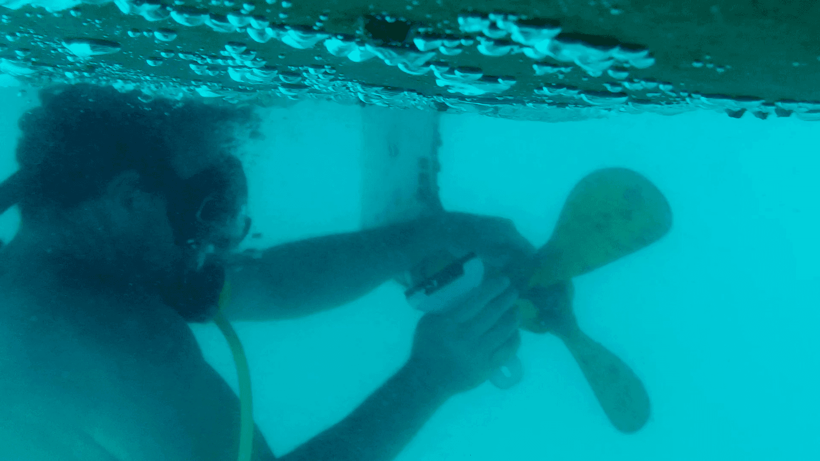 Person underwater using a scraperite tool on a boat's propeller.